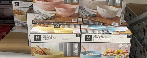 A stack of ceramic prep and serve bowls in various colors (pink, blue, and beige) inside their packaging displayed on a shelf.