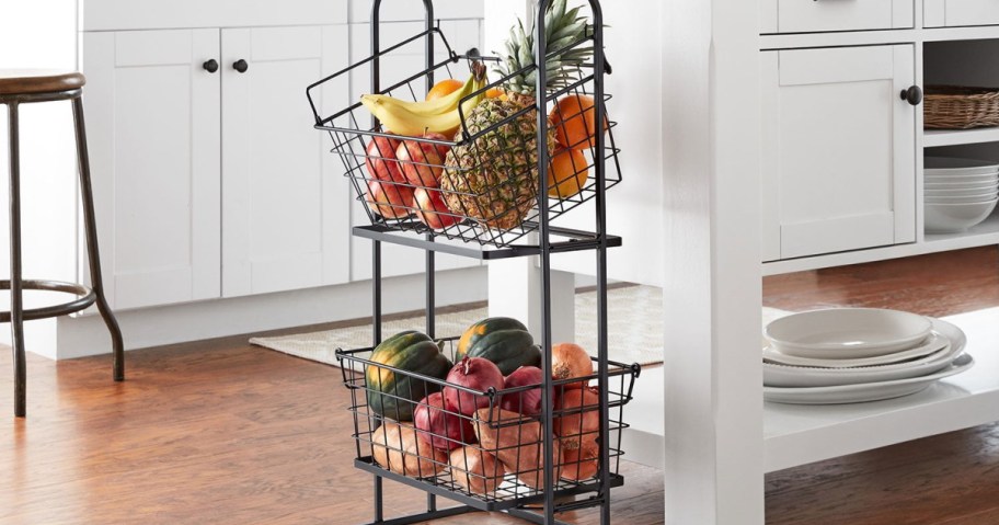 Two-tiered wire basket stand in a kitchen holds fresh fruit including bananas, pineapples, and apples.