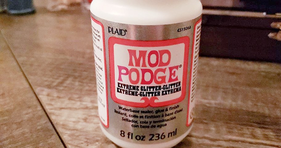 A bottle of Mod Podge Extreme Glitter stands on a wooden table. The label is white and pink, stating it is an 8 fl oz waterbase sealer, glue, and finish.