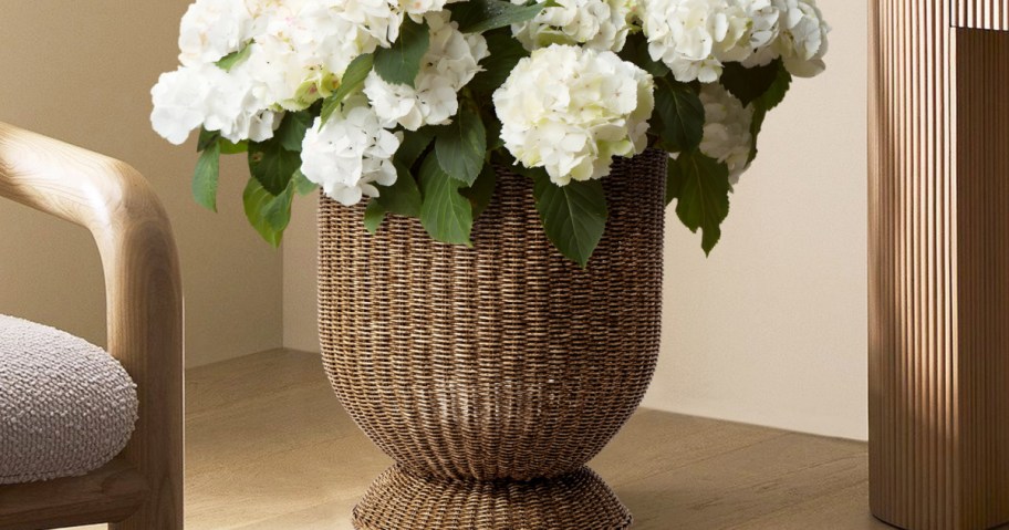 A woven wicker vase holds lush white hydrangeas with green leaves, placed on a light wooden floor.