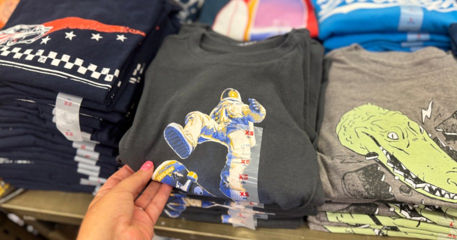 A hand holds a dark t-shirt featuring a colorful astronaut graphic. Nearby, folded shirts display designs of crocodiles and race-themed patterns. XS size tags are visible.
