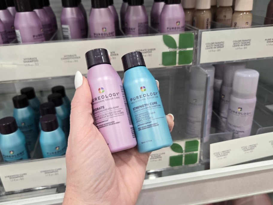 A hand holds two Pureology shampoo bottles, one purple "Hydrate" and one blue "Strength Cure," in front of a store shelf filled with similar products.