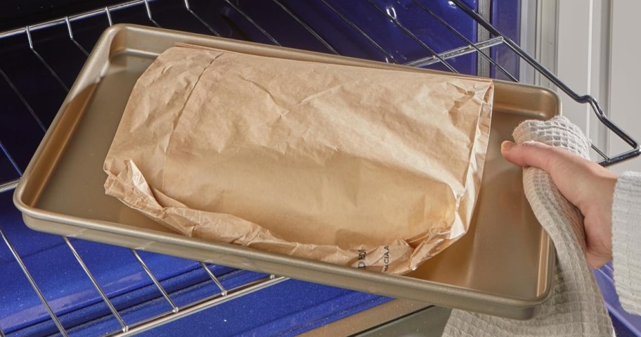 A baking tray holding a brown parchment paper cooking bag is being placed into an oven. A hand holding a dishcloth carefully supports the tray.