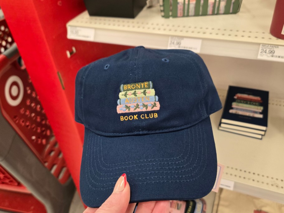 Blue baseball cap with embroidered text "Book Club" below stacked books labeled "Bronte" and "Austen." Displayed in a retail setting with books in the background.