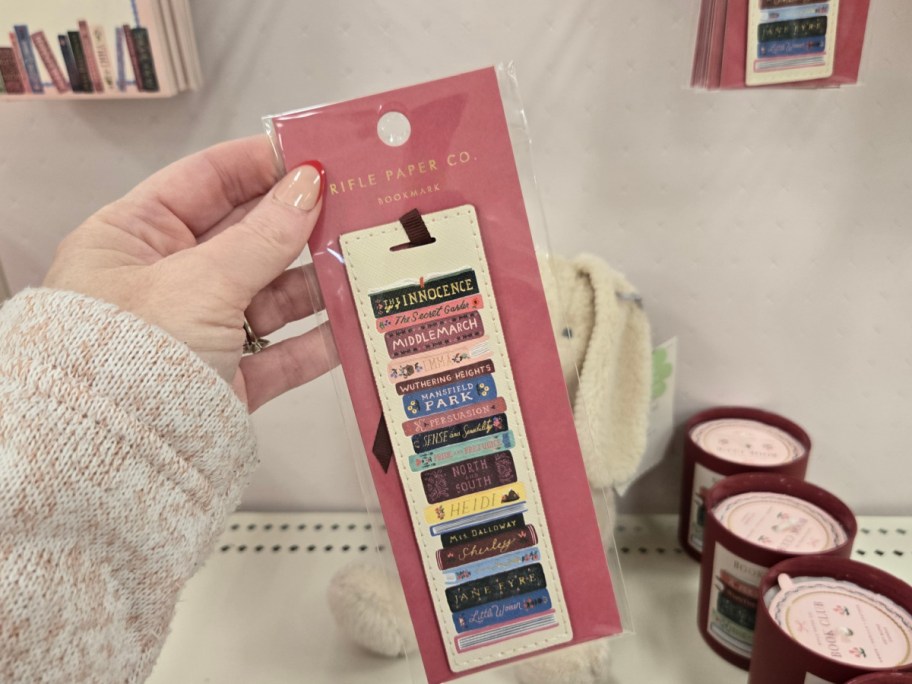 A hand holds a Rifle Paper Co. bookmark featuring colorful book spines, titles include classics like "Wuthering Heights." 