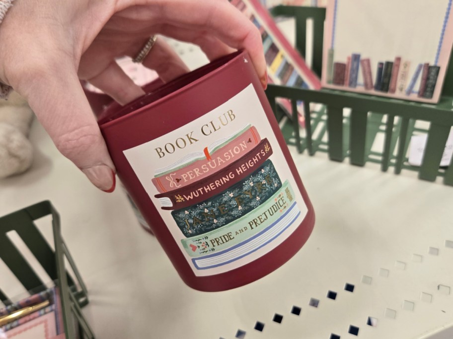 A hand holds a red candle labeled "Book Club," featuring illustrated book spines with titles like "Persuasion" and "Pride and Prejudice." Bookshelves are visible in the background.