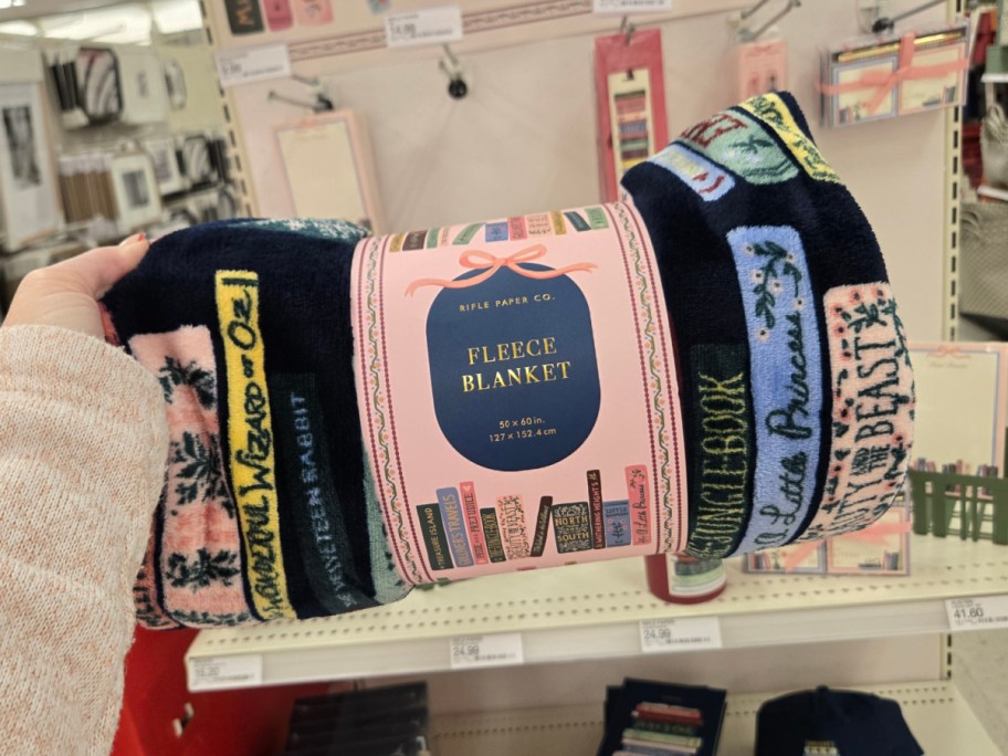 A hand holds a rolled fleece blanket featuring colorful book spines with classic titles.