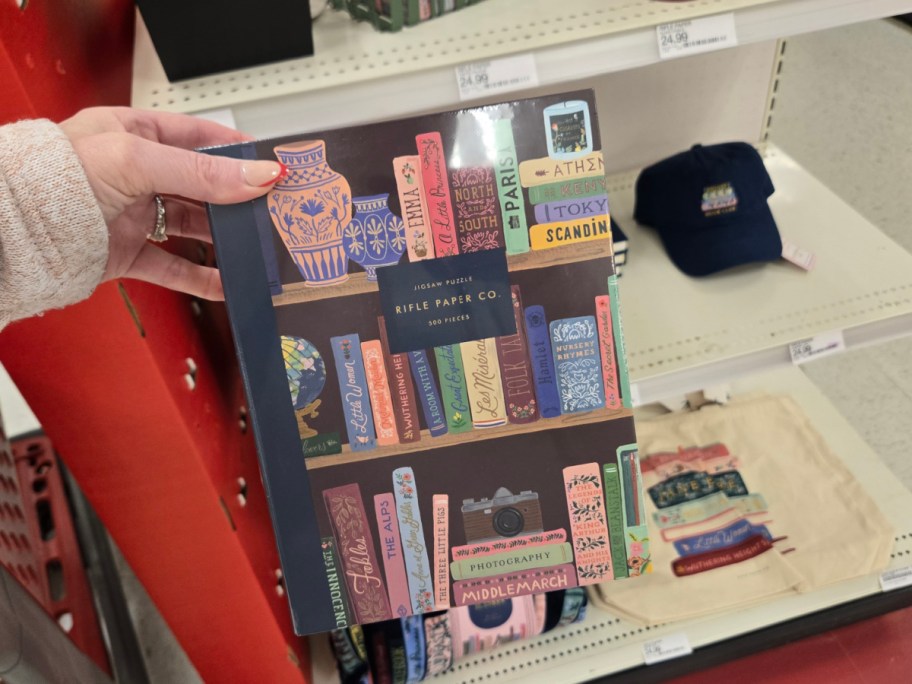 A person's hand holds a Rifle Paper Co. jigsaw puzzle depicting colorful books and vases on a shelf. Nearby, a blue cap and canvas tote are displayed.