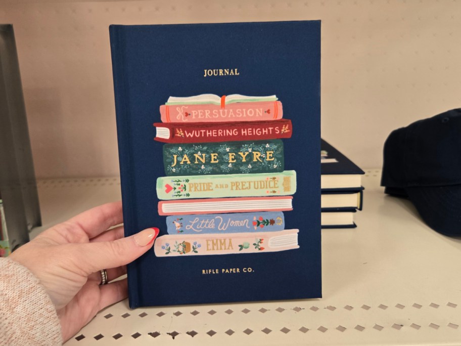 A hand holds a blue journal with illustrations of classic novels stacked: "Persuasion," "Wuthering Heights," "Jane Eyre," "Pride and Prejudice," "Little Women," and "Emma." The cover is colorful.