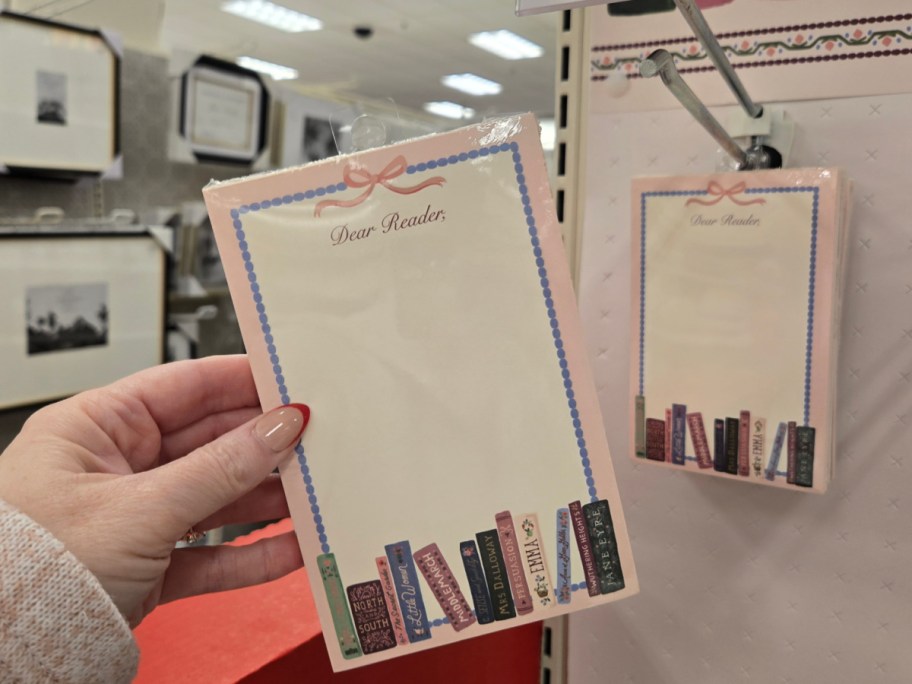 A hand holds a decorative notepad with "Dear Reader" at the top and colorful book spines at the bottom. A similar notepad is displayed on a store rack.