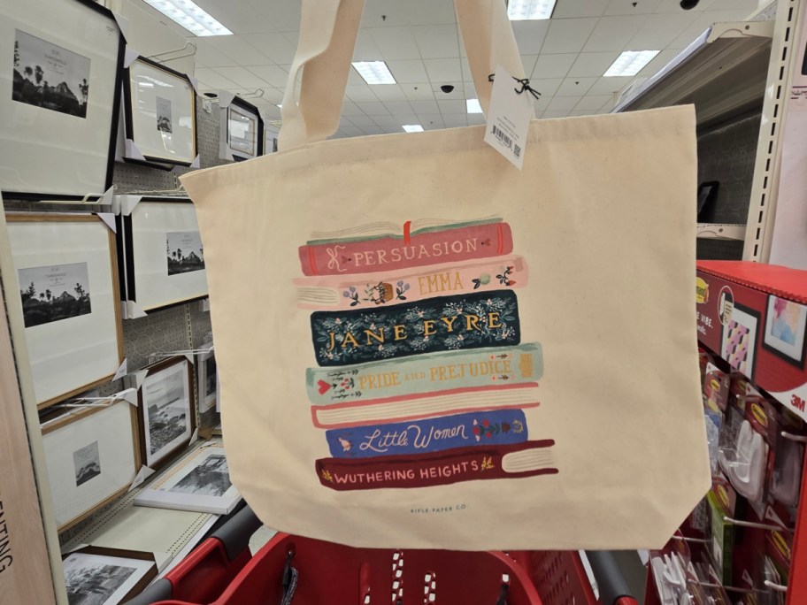 Canvas tote bag with colorful illustrations of classic novels stacked: "Persuasion," "Emma," "Jane Eyre," "Pride and Prejudice," "Little Women," and "Wuthering Heights." Inside a store with framed art on shelves.