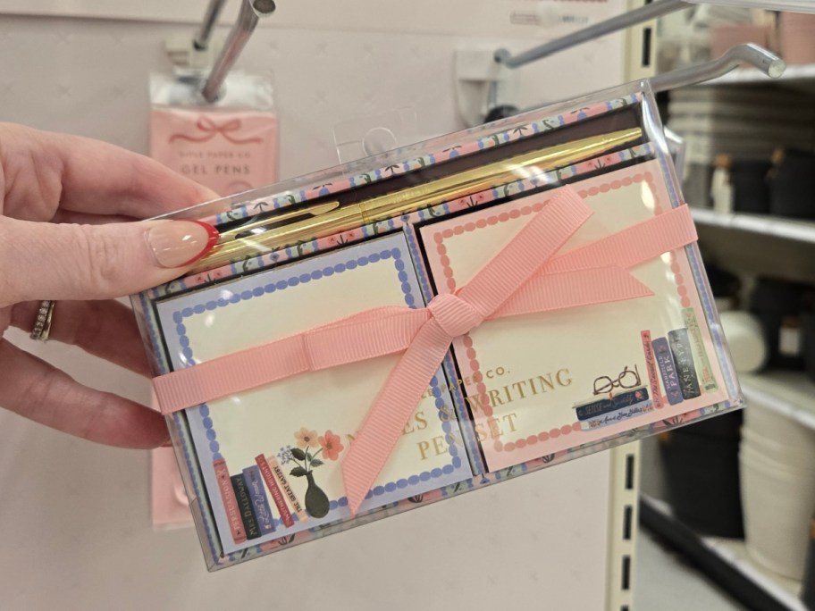 A hand holds a decorative note and pen set with a pink ribbon. Store shelves are visible in the background.
