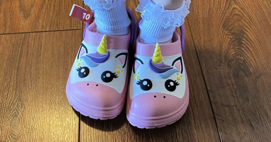 A pair of pink children's clogs featuring a unicorn face and a yellow horn, worn with white ruffled socks.