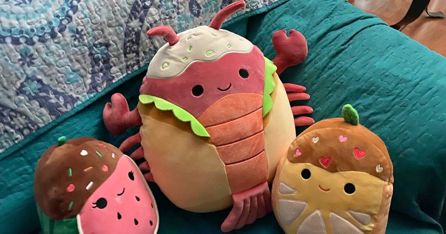 Three smiling Squishmallows plush toys resembling food including a lobster roll sit on a teal quilted sofa. 