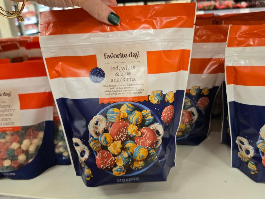 Close-up of a hand holding a bag of "Favorite Day" red, white, and blue snack mix. The package features colorful treats and a festive design.