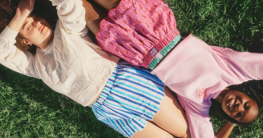 Two people lie on green grass in colorful shorts and pastel tops, basking in sunlight.