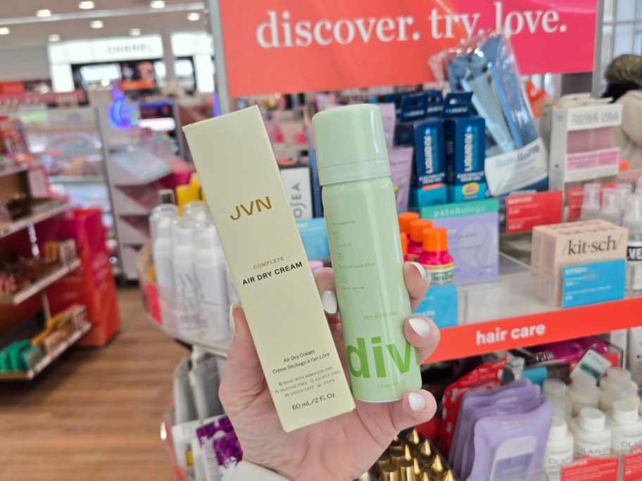 A person holds JVN Air Dry Cream and a green bottle of dry shampoo in a store's hair care aisle. Background shows colorful product displays.