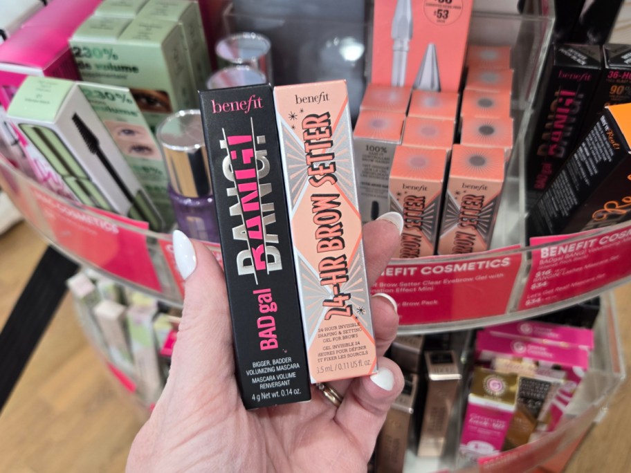 A hand holds a Benefit BADgal BANG! mascara and 24-HR Brow Setter. The background shows a colorful makeup display, creating a vibrant retail atmosphere.