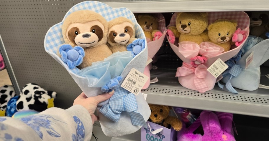 hand holding a plush bouquet with a mama and baby sloth, more bouquets with bears on a shelf behind it