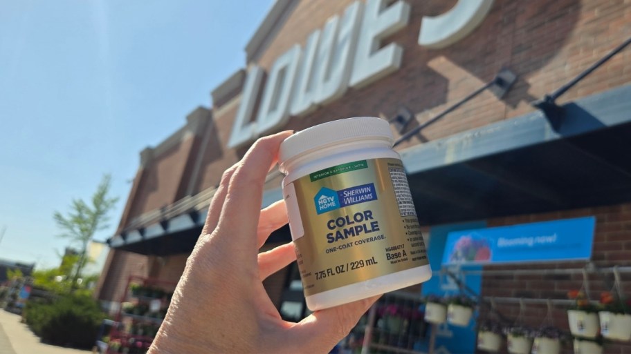 womans hand holding a paint sample jar up in front of a lowes store