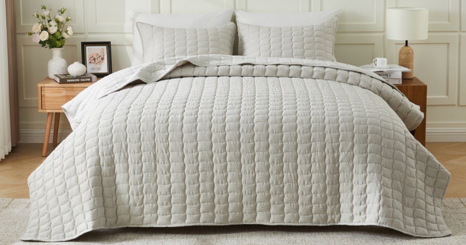 A neatly made bed with a textured, light gray quilt and matching pillows. A wooden nightstand holds flowers and a framed photo.