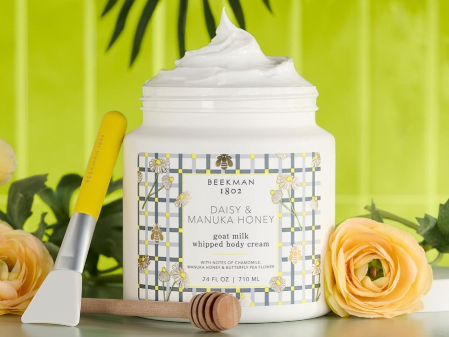 A jar of Beekman 1802 Daisy & Manuka Honey goat milk whipped body cream with a floral design, surrounded by yellow roses.