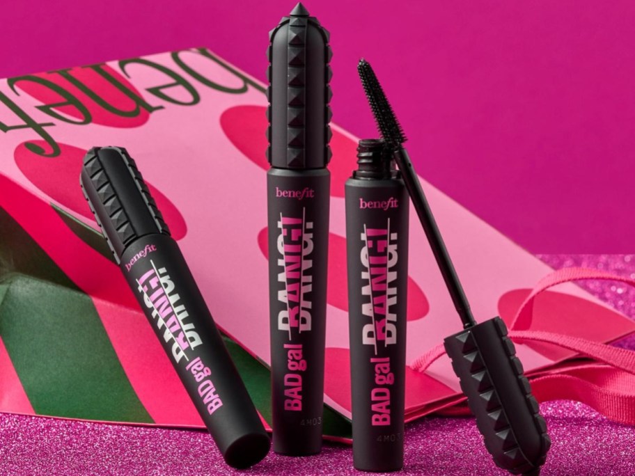 Mascara tubes on a pink glittery surface, with bold “BADgal BANG!” labeling. A pink and red heart-patterned bag is in the background.