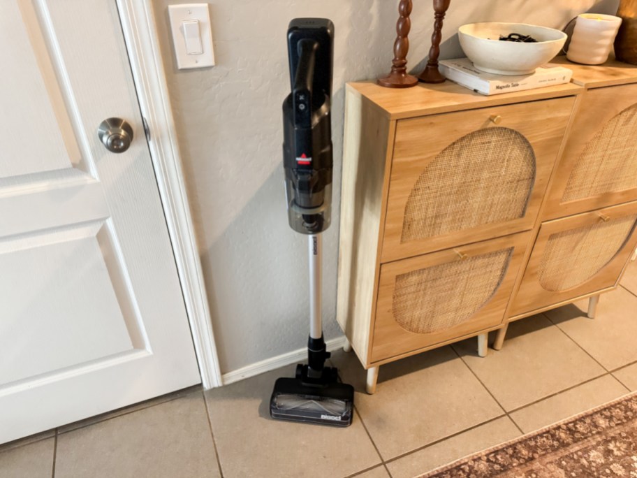 A cordless vacuum cleaner stands upright next to a light-colored wooden cabinet with four basket weave doors.
