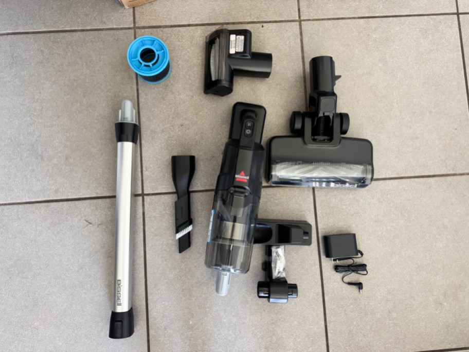 Vacuum cleaners, brush heads, tubes, filters, adapters, small tools and other parts are lined up on a tiled floor.