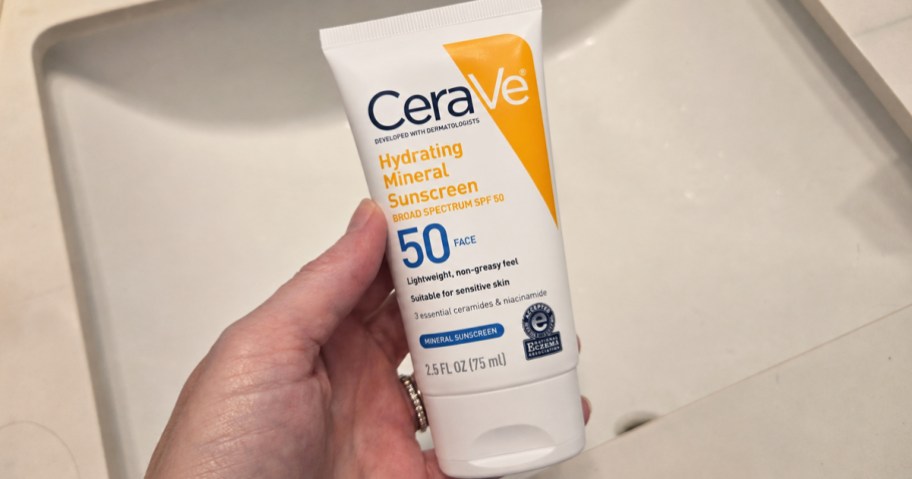 hand holding a tube of CeraVe Mineral SPF 50 Face Sunscreen with bathroom sink in background