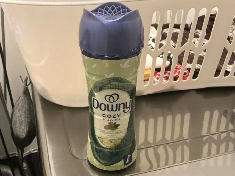A bottle of Downy Cozy Collection scent beads on a dryer with a basket of clean clothes behind it