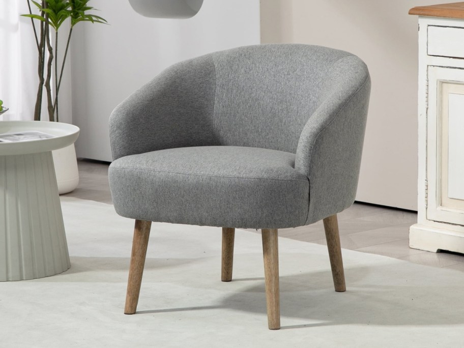 Grey upholstered armchair with wooden legs on a light rug in a cozy room. Beside it, a white side table holding magazines.
