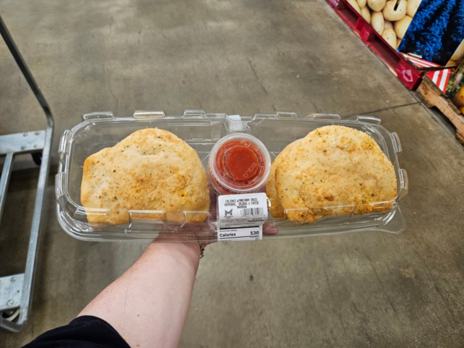 Two calzones with a side of dipping sauce in a plastic container held by a hand.