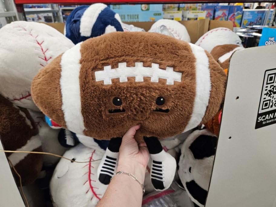 A plush football toy with a cute face being held by a person in a store among other sports-themed plush toys.