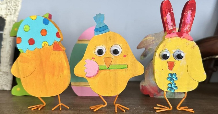 Three colorful wooden chicks with large eyes are on display. Each has distinct decorations: a polka-dot egg, a hat with a flower, and red bunny ears.