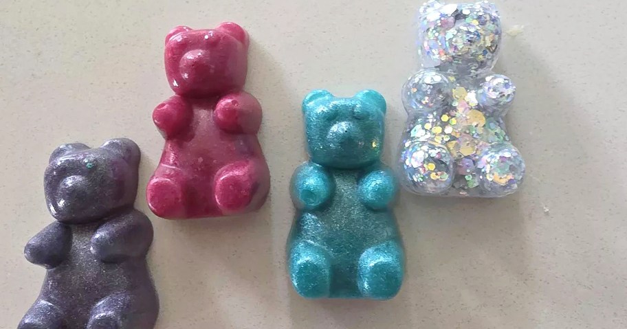 Four glittery bear-shaped crayons in different colors&mdash;purple, pink, teal, and silver with sparkles&mdash;are lined up on a light surface.