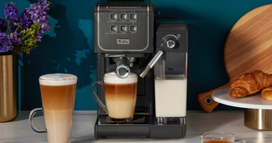 A sleek espresso machine froths milk into a glass cup of coffee beside a wooden tray with croissants.