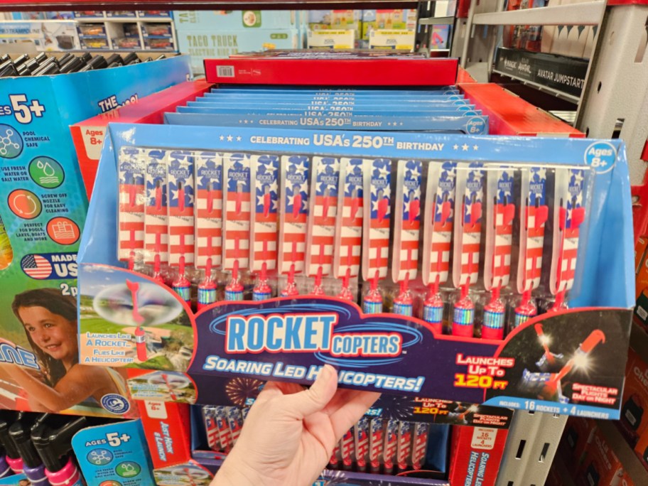 A box of "Rocket Copters" fireworks featuring red, white, and blue designs celebrating the USA's 250th birthday, with a person’s hand holding the box in a store aisle.