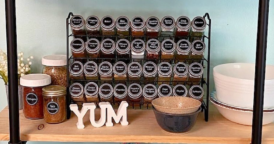 A spice rack filled with labeled jars of various spices is set on a wooden shelf. Below, the word "YUM" in white letters, with bowls and jars nearby.