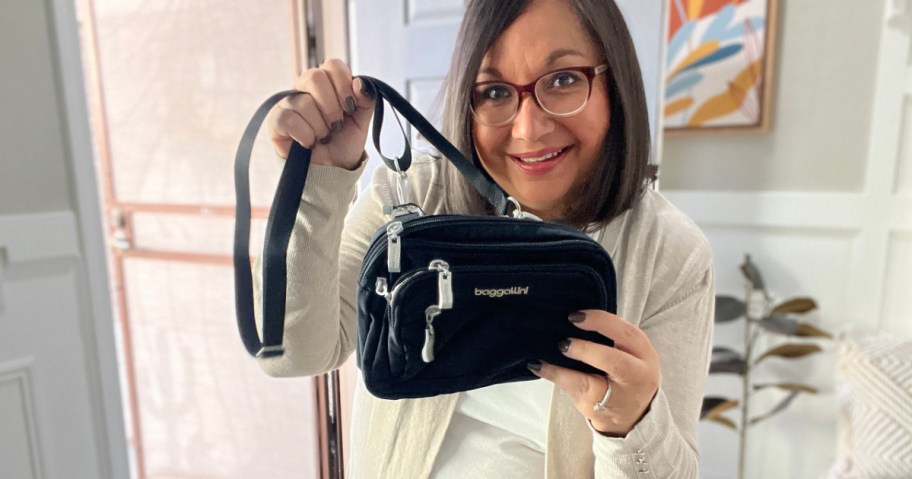 A cheerful woman with glasses holds up a small black bag with silver zippers. She's in a bright room adorned with modern art and a plant.