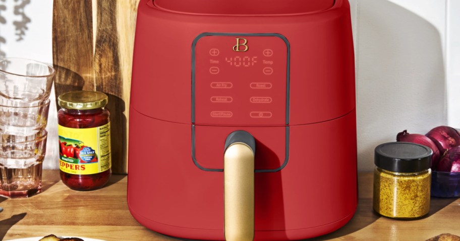 Red air fryer with a gold handle on a wooden counter, surrounded by glass cups, a jar of peppers, onions, and a spice jar.