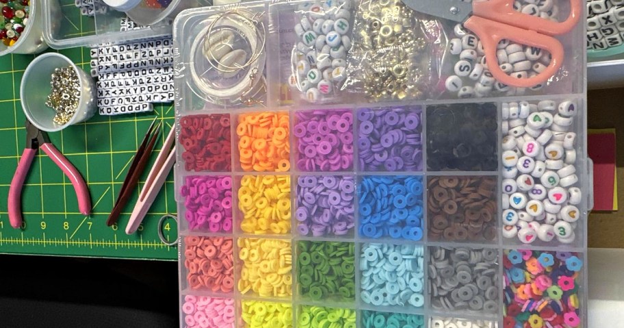 A colorful craft box filled with beads sorted by color, surrounded by crafting tools like scissors and pliers on a cutting mat.
