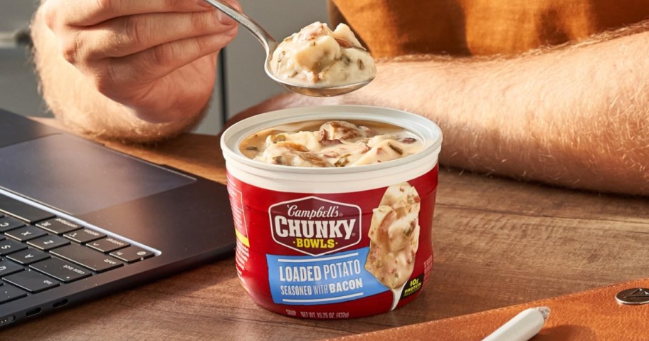 A person holds a spoon of creamy soup over a Campbell&rsquo;s Chunky Loaded Potato Bowl, with a laptop and brown table setting in the background.