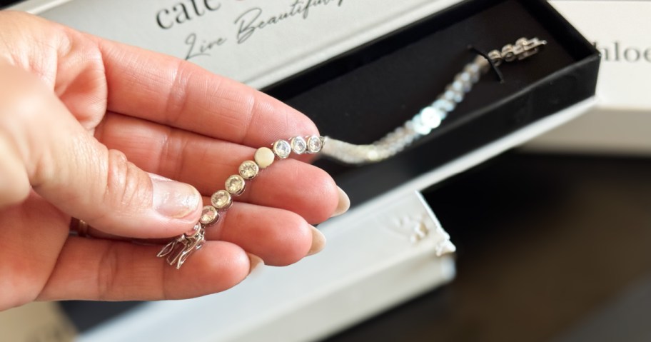 A hand holds a sparkling silver bracelet with round gemstones, above an open jewelry box labeled "Live Beautifully."