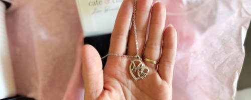 A hand holding a silver necklace with a heart pendant engraved with "Mom." The background shows a Cate & Chloe jewelry box on soft pink tissue paper.