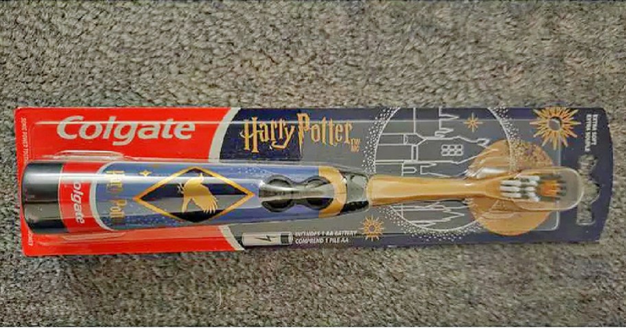 Colgate Harry Potter electric toothbrush in colorful packaging. Features a blue handle with a golden snitch design. Set against a textured gray background.