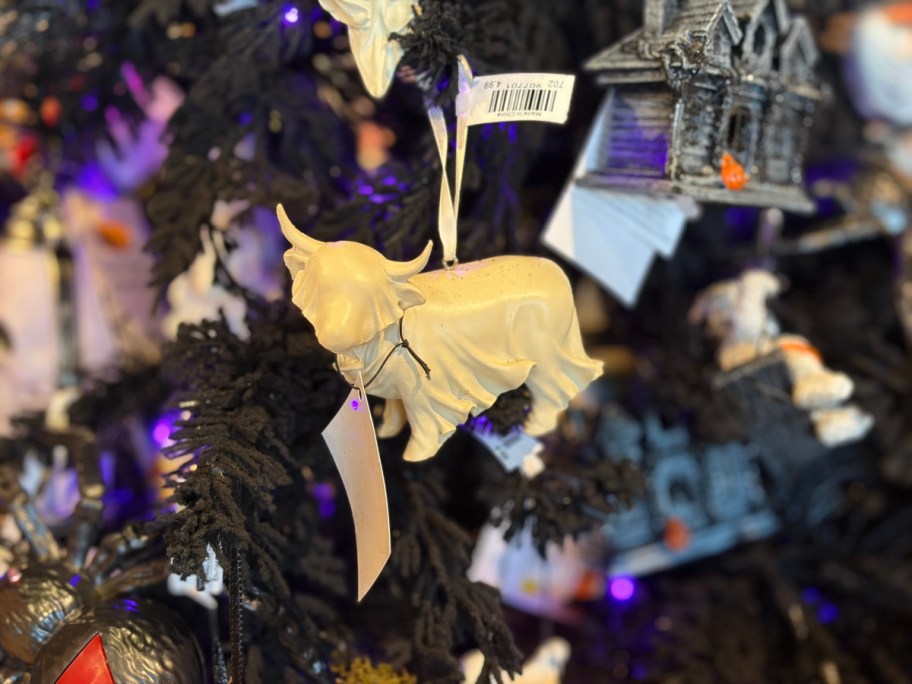 A cream-colored cow ornament hanging on a black Christmas tree adorned with various decorations.