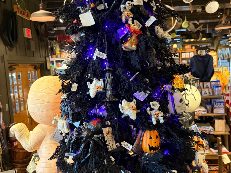 A black Halloween-themed tree decorated with various ornaments, including ghosts, a pumpkin, and other spooky decorations, set in a store filled with holiday items.