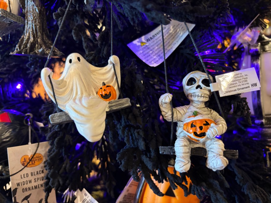 Two Halloween ornaments: a ghost swinging on a swing holding a pumpkin, and a mummy on another swing also holding a pumpkin, hanging on a black tree.