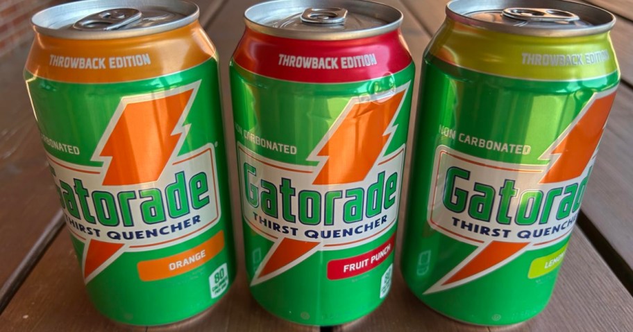 Three Gatorade Thirst Quencher cans labeled as "Throwback Edition" with flavors: Orange, Fruit Punch, and Lemon-Lime, set on a wooden table.